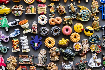 A colorful assortment of decorative fridge magnets depicting various objects such as donuts, animals, owls, fruits, and Italian-themed items. The magnets are arranged closely together on a dark background, showcasing a wide variety of shapes and designs, including smiley faces and motorcycles.