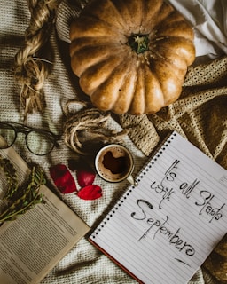 An autumn-themed flat lay featuring a large pumpkin on a textured fabric background. Nearby, an open book and a spiral notebook with handwritten text are visible, along with reading glasses, red rose petals, a small cup of coffee, and a sprig of greenery.