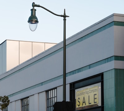 A modern building with a rectangular shape, featuring horizontal lines of teal and white. There is a prominent street lamp with a curved arm extending above the building, and a sign that reads 'Liquidation Rug Company SALE' in yellow and black on one of the building walls. The top part of the building has reflective glass windows with a soft beige tint. There are also narrow sections of vertical windows below the main signage.