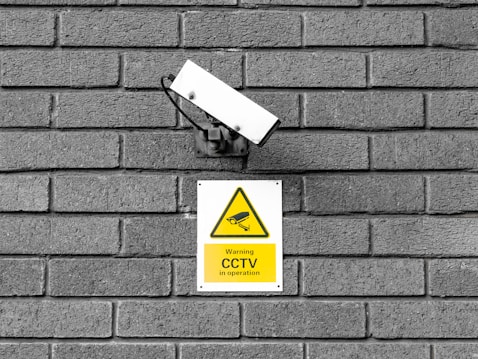 A CCTV surveillance camera is mounted on a gray brick wall. Below the camera is a yellow warning sign indicating that CCTV is in operation with an image of a camera in a triangular warning symbol.