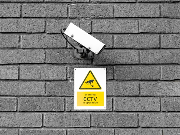 A CCTV surveillance camera is mounted on a gray brick wall. Below the camera is a yellow warning sign indicating that CCTV is in operation with an image of a camera in a triangular warning symbol.