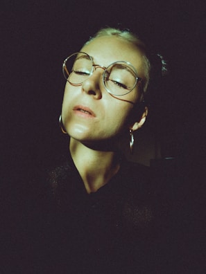 A person with a serene expression and closed eyes is illuminated against a dark background. The individual wears large, round eyeglasses and hoop earrings. There is a strong contrast between the highlights on the face and the surrounding darkness.