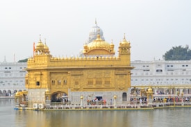 A magnificent structure with ornate golden walls and domes stands over a body of water, surrounded by a crowd of people. The architecture features intricate carvings and decorative elements, and a larger white building can be seen in the background.