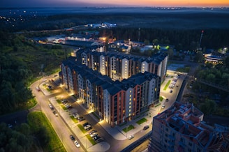 Aerial view of a modern apartment complex illuminated at dusk. The well-lit buildings have a mix of dark and light exterior tones. Surrounding the complex are neatly arranged parking lots with cars. In the background, there are patches of green areas and distant city lights under a darkening sky.