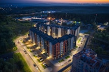 Aerial view of a modern apartment complex illuminated at dusk. The well-lit buildings have a mix of dark and light exterior tones. Surrounding the complex are neatly arranged parking lots with cars. In the background, there are patches of green areas and distant city lights under a darkening sky.