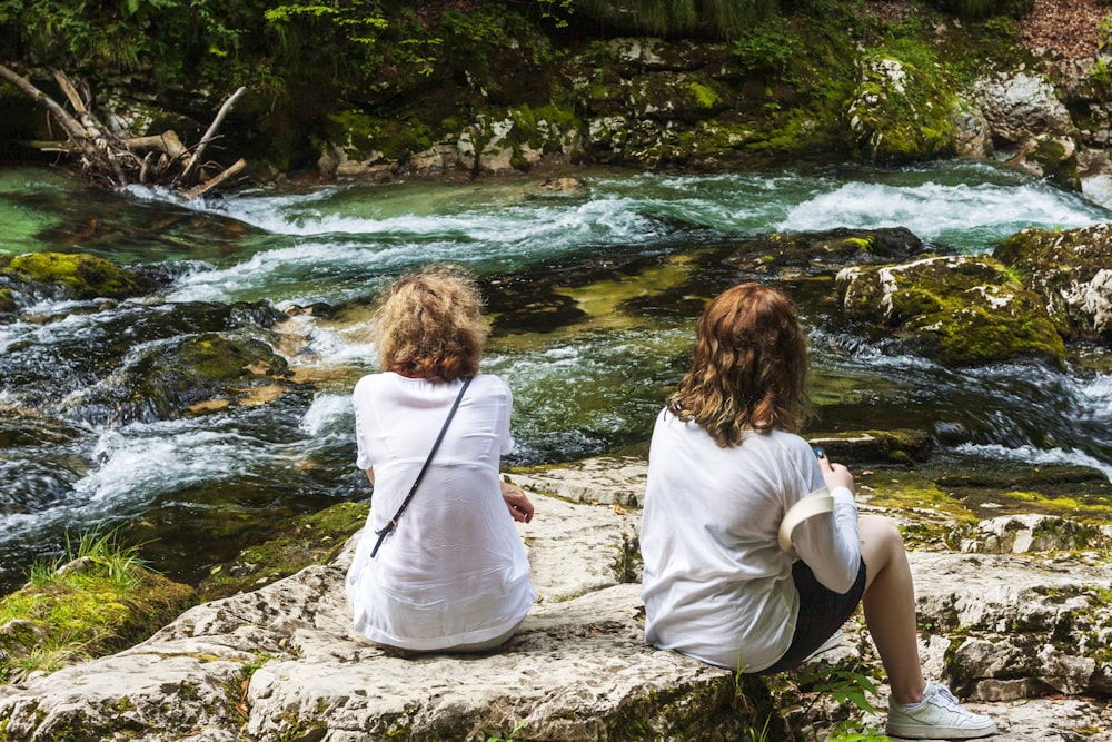 2 women sitting on rock near river during daytime