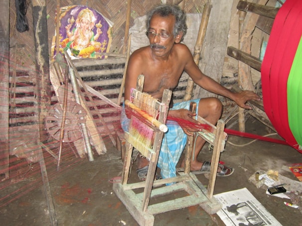 An older man with glasses is seated and working with a weaving loom in an indoor setting. He is wearing a traditional wrap around his waist and appears focused on his task. Beside him, there is a vivid display of colored threads. Behind him, a poster of a deity suggests a cultural or religious significance. The room contains various tools and objects related to weaving.