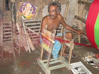 An older man with glasses is seated and working with a weaving loom in an indoor setting. He is wearing a traditional wrap around his waist and appears focused on his task. Beside him, there is a vivid display of colored threads. Behind him, a poster of a deity suggests a cultural or religious significance. The room contains various tools and objects related to weaving.