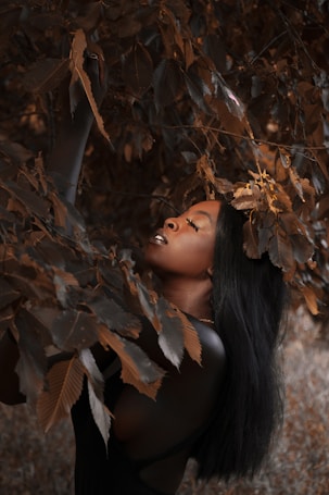 A person with closed eyes is surrounded by dense, dark foliage, creating a serene atmosphere. The leaves have a rich, brownish color contrasting with the smooth texture of the individual's skin. The composition gives a sense of being intertwined with nature.