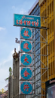 A series of vertical blue signs with red text displayed on the side of a building, featuring Chinese characters. The building has a modern architectural design with glass windows and a distinctive yellow facade. There are ornamental features and some greenery visible in the vicinity.