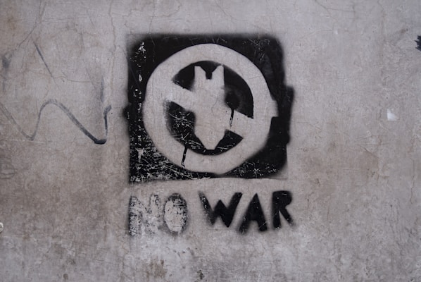 A stencil graffiti on a textured wall features a circle with a plane inside it, crossed out diagonally, symbolizing anti-war sentiment. Below this symbol, the words 'NO WAR' are spray-painted in bold, black letters.