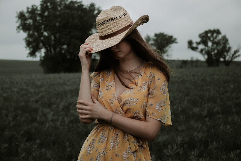 黄色と白の花柄のドレスを着た女性が、緑の草原に立って茶色の麦わら帽子をかぶっています