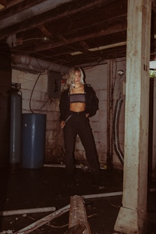 A person with long hair stands in a dimly lit basement. The surroundings are industrial, with exposed pipes, a blue cylindrical tank, and concrete walls. The individual is wearing a black jacket and pants, with a striped top underneath.