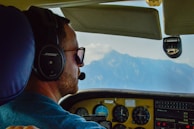 A pilot is wearing a headset and sunglasses inside a cockpit, focusing on flying. The cockpit dashboard displays various instruments and gauges. Through the window, a clear view of distant mountains under a blue sky is visible.