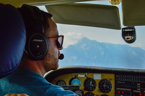 A pilot is wearing a headset and sunglasses inside a cockpit, focusing on flying. The cockpit dashboard displays various instruments and gauges. Through the window, a clear view of distant mountains under a blue sky is visible.