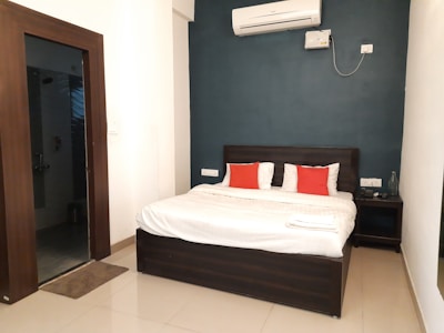 A modern bedroom features a neatly made double bed with white linens and two red accent pillows. The bed is positioned against a dark blue accent wall. Above the bed, an air conditioning unit is mounted. A bedside table to the right holds a glass bottle and a telephone. The floor is tiled in a light beige color, and an open door on the left leads to what appears to be a bathroom.