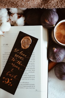 An open book with a page marked by a decorative bookmark that reads 'I love books to the moon and back'. The bookmark features a gold wax seal and celestial designs. Surroundings include ripe figs, a cup of coffee, and soft cotton plants, set on a cozy, textured surface.