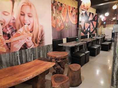 A cozy dining area features rustic wooden tables and stools combined with modern, black cushioned seating. The walls are adorned with large, vibrant images of people enjoying pizza and artistic renderings of pizza slices. Ambient lighting with multiple hanging spherical lights adds warmth and an inviting atmosphere.