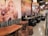 A cozy dining area features rustic wooden tables and stools combined with modern, black cushioned seating. The walls are adorned with large, vibrant images of people enjoying pizza and artistic renderings of pizza slices. Ambient lighting with multiple hanging spherical lights adds warmth and an inviting atmosphere.