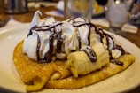 A plate of crepes topped with whipped cream, sliced bananas, chocolate sauce, and a light dusting of powdered sugar. The focus is on the dessert which is placed on a table with blurred background elements.