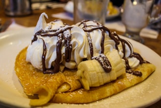 A plate of crepes topped with whipped cream, sliced bananas, chocolate sauce, and a light dusting of powdered sugar. The focus is on the dessert which is placed on a table with blurred background elements.