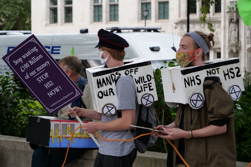 Two people are participating in a protest, wearing cardboard costumes with slogans related to environmental conservation. One of them holds a sign with a message against HS2, a high-speed railway project. They are wearing face masks, and there is a police van in the background.