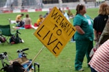 People gathered in a park with signs, including a prominent yellow sign that reads 'WE WANT TO LIVE'. Individuals are seated on the grass, some in casual clothing and others in medical scrubs, suggesting a protest or public demonstration.