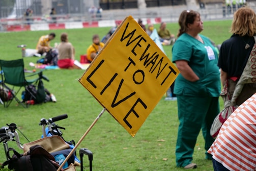 People gathered in a park with signs, including a prominent yellow sign that reads 'WE WANT TO LIVE'. Individuals are seated on the grass, some in casual clothing and others in medical scrubs, suggesting a protest or public demonstration.