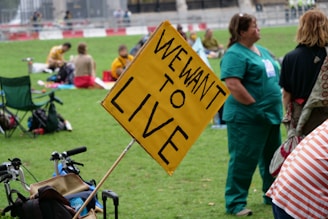 People gathered in a park with signs, including a prominent yellow sign that reads 'WE WANT TO LIVE'. Individuals are seated on the grass, some in casual clothing and others in medical scrubs, suggesting a protest or public demonstration.