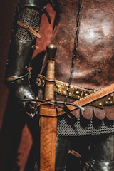 A close-up view of a medieval-style armor with intricately designed metal plates and chainmail. The leather belt features metal studs and a sword with an ornate, leather-wrapped handle rests at the side.