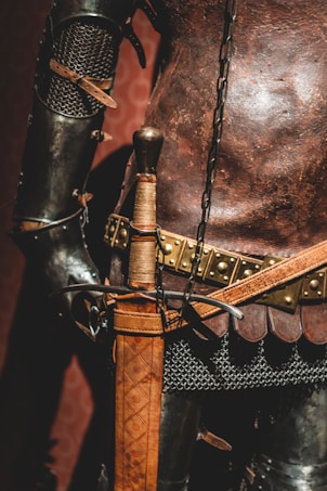 A close-up view of a medieval-style armor with intricately designed metal plates and chainmail. The leather belt features metal studs and a sword with an ornate, leather-wrapped handle rests at the side.