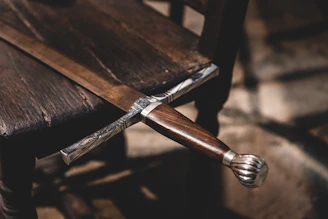 A close-up of a finely crafted sword resting on a wooden table with leather-bound armors in the background.