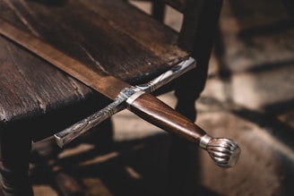 An intricately designed sword with a wooden handle is laid across a wooden chair. The sword's hilt is detailed with metal craftsmanship. The setting has a rustic and historical feel, with shadows cast by natural light.