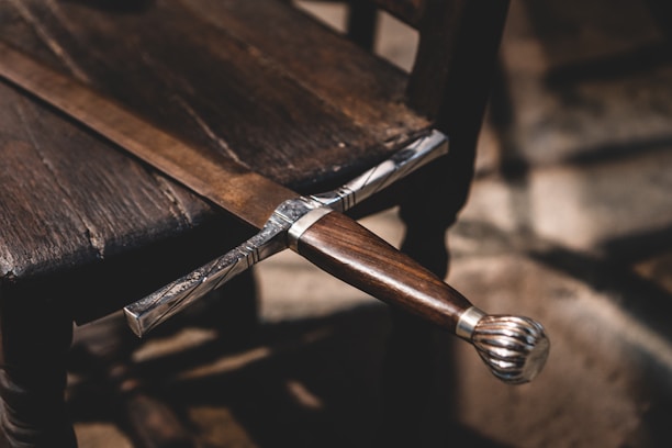 An intricately designed sword with a wooden handle is laid across a wooden chair. The sword's hilt is detailed with metal craftsmanship. The setting has a rustic and historical feel, with shadows cast by natural light.