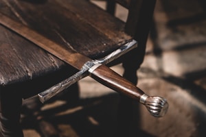 An intricately designed sword with a wooden handle is laid across a wooden chair. The sword's hilt is detailed with metal craftsmanship. The setting has a rustic and historical feel, with shadows cast by natural light.