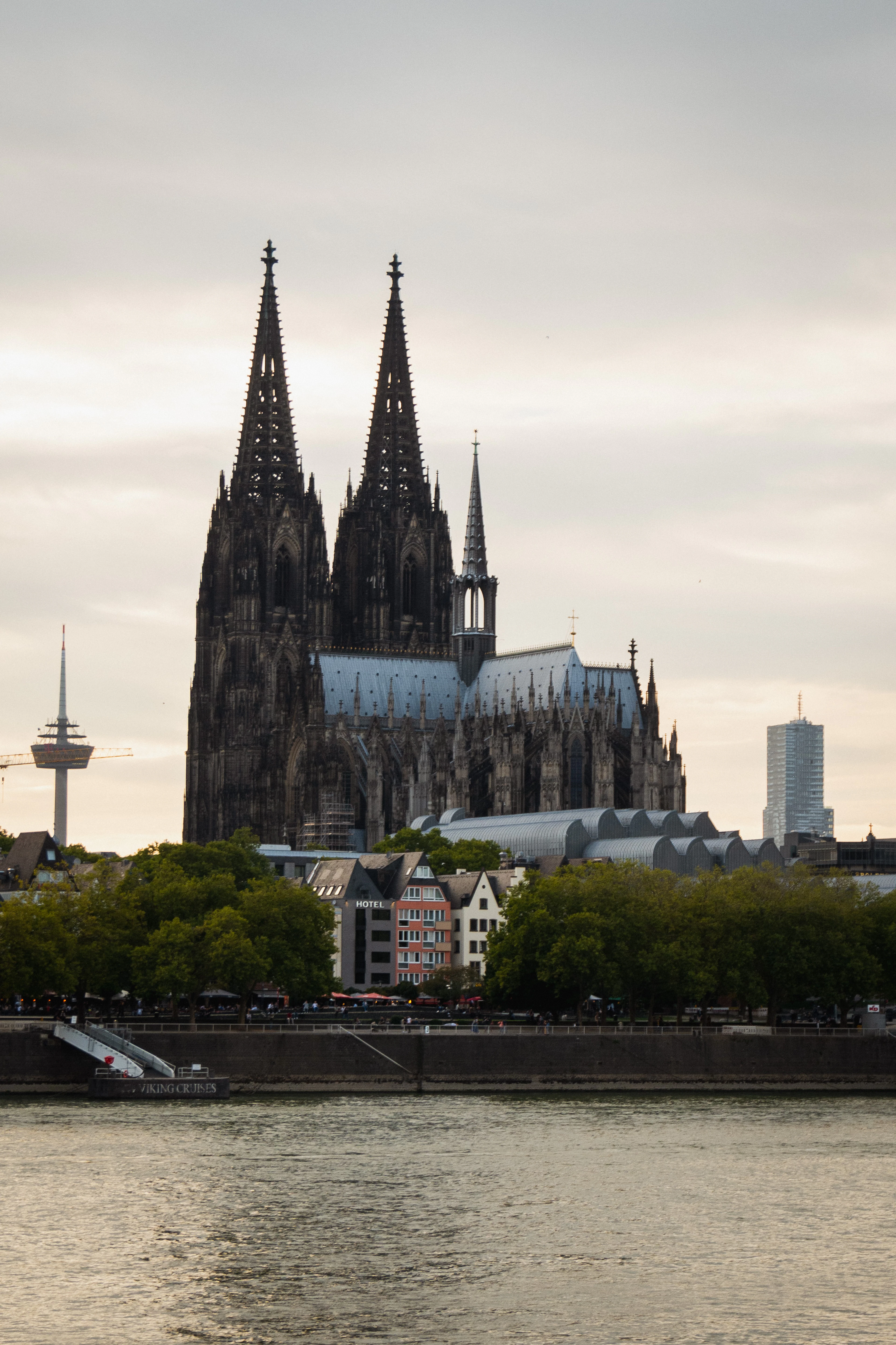 Cologne Cathedral's silhouette rises over the Rhine at dusk beside colorful old-town houses and a river boat.
