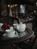 A close-up of a white teapot and teacup set on a glass-topped wicker table, with folded red and brown napkins next to them. The background features a blurred outdoor setting with soft lighting.