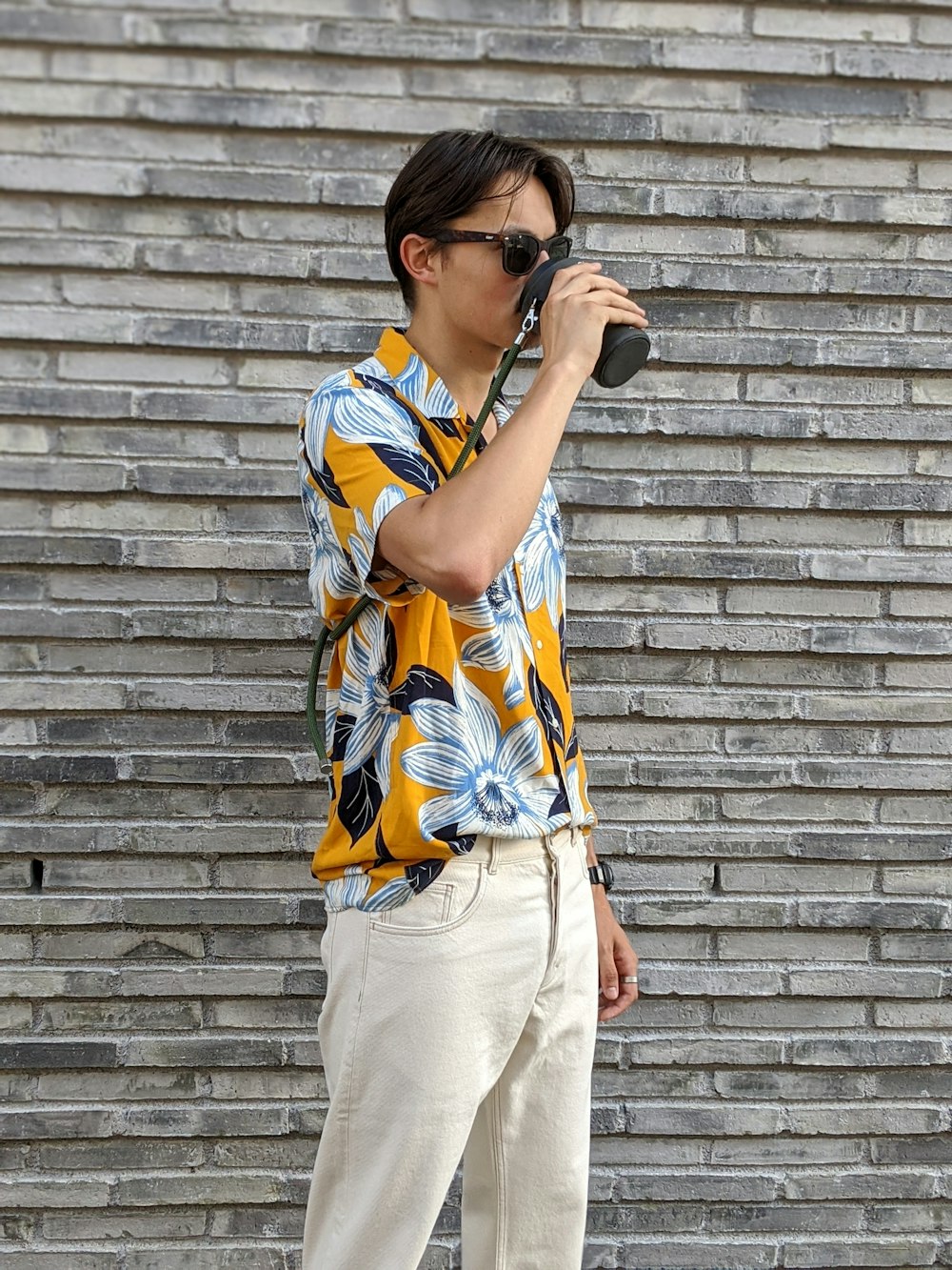 woman in yellow white and red floral shirt and beige pants standing beside brick wall