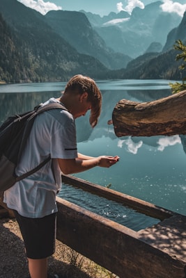 A person with short hair is standing beside a wooden water trough, collecting water with their hands. They are wearing a backpack and a white shirt. The background features a serene lake surrounded by mountains, with a clear, sunny sky reflecting on the water's surface.