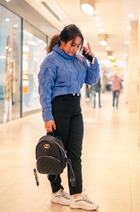 A person in a blue sweater is standing in a brightly lit indoor shopping mall. They are holding a phone up to their ear and carrying a black backpack. The person is wearing black pants and white sneakers. There are blurred figures and shop displays in the background.