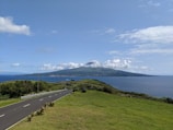 A scenic view of a paved road running alongside green fields, leading to the distant ocean. A mountainous island sits in the background under a partly cloudy sky.