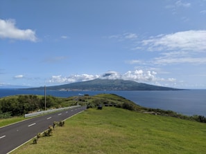 A scenic view of a paved road running alongside green fields, leading to the distant ocean. A mountainous island sits in the background under a partly cloudy sky.