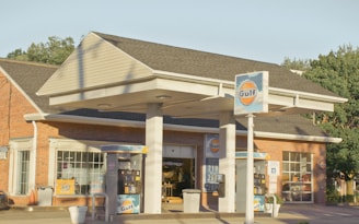 A gas station with Gulf branding, featuring two fuel pumps beneath a canopy. The brick building in the background has large windows. There are signs with fuel prices and various advertisements. The scene is lit by warm, natural sunlight.