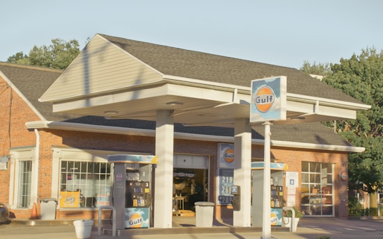 A gas station with Gulf branding, featuring two fuel pumps beneath a canopy. The brick building in the background has large windows. There are signs with fuel prices and various advertisements. The scene is lit by warm, natural sunlight.
