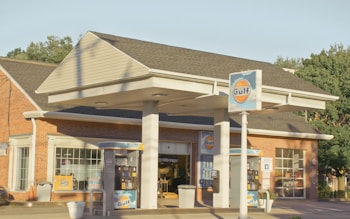 A gas station with Gulf branding, featuring two fuel pumps beneath a canopy. The brick building in the background has large windows. There are signs with fuel prices and various advertisements. The scene is lit by warm, natural sunlight.