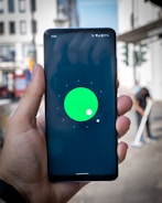 A hand holding a smartphone with a display showing a green circle with a smaller white circle on a black background. The screen also displays the time 11:32, Wi-Fi signal, battery percentage, and other notification icons at the top. The background appears to be an outdoor urban setting, slightly blurred, with buildings and a person visible.