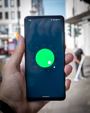 A hand holding a smartphone with a display showing a green circle with a smaller white circle on a black background. The screen also displays the time 11:32, Wi-Fi signal, battery percentage, and other notification icons at the top. The background appears to be an outdoor urban setting, slightly blurred, with buildings and a person visible.