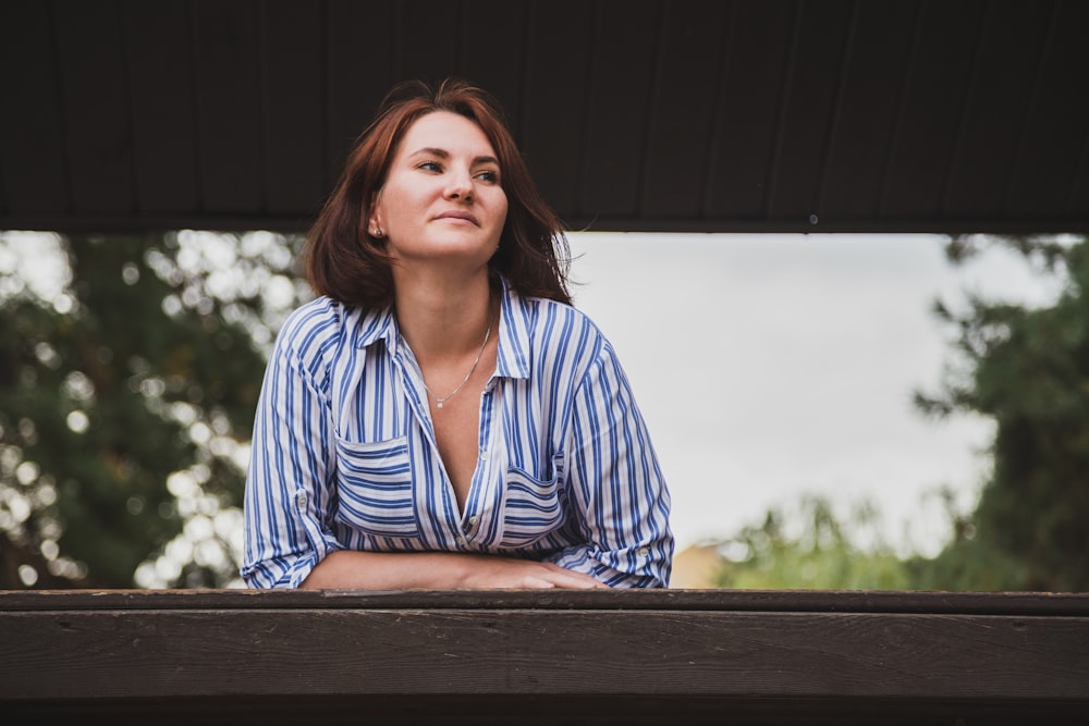 woman in blue and white striped dress shirt sitting on brown wooden bench during daytime