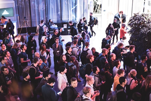 A large group of people gathered in an indoor setting, seemingly attending a public event or conference. The crowd consists of individuals in casual attire, many of whom are holding drinks or using their phones. The background features large windows, a tree, and a digital display screen.