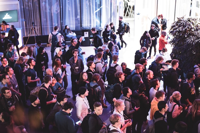 A large group of people gathered in an indoor setting, seemingly attending a public event or conference. The crowd consists of individuals in casual attire, many of whom are holding drinks or using their phones. The background features large windows, a tree, and a digital display screen.